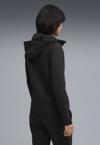 Haut à capuche noir avec une finition texturée, présentant un design ajusté, une capuche réglable et des poches latérales. Tissu lisse et chaud, adapté pour être superposé.