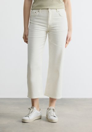 Άτομο που φοράει εκρού καλλιεργημένα τζιν με φαρδιά πόδια και λευκά αθλητικά παπούτσια, στέκεται σε ανοιχτό γκρι πάτωμα μπροστά σε απλό λευκό φόντο.