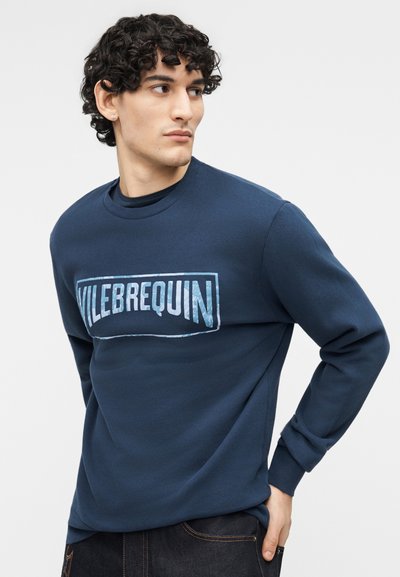 Νέος άνδρας με σγουρά μαλλιά που φοράει φούτερ Vilebrequin σε ναυτικό μπλε χρώμα, κοιτάζοντας προς τα πλάγια με το ένα χέρι στην τσέπη του.