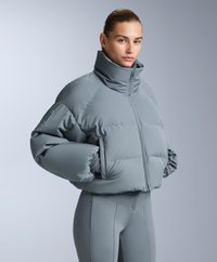 Pufasta siva jakna s visokim ovratnikom, prednjim patentnim zatvaračem i previše velikim rukavima; uparena s uskim sivim hlačama. Glatka, mat tekstura.