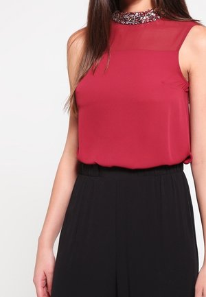 Hihaton punainen pusero, jossa on läpikuultava yläosa ja helmin koristeltu kaulus, yhdistetty korkeavyötäröisiin mustiin, leveälahkeisiin housuihin naisella.