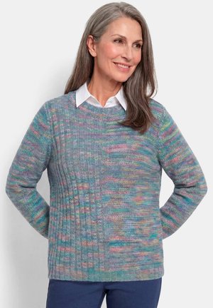 Femme d'âge moyen souriante aux longs cheveux gris porte un pull en tricot multicolore par-dessus une chemise blanche à col et un pantalon foncé sur un fond uni.