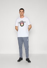 Hvid bomulds t-shirt med et farverigt "Tyskland" grafik og sorte accenter, parret med grå sweatpants med hvide striber og et Adidas-logo.