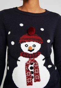 Maglione lavorato a maglia blu navy con un design di un pupazzo di neve, dotato di un cappello rosso, pompon e sciarpa, fiocchi di neve bianchi e una sciarpa a maglia testurizzata.