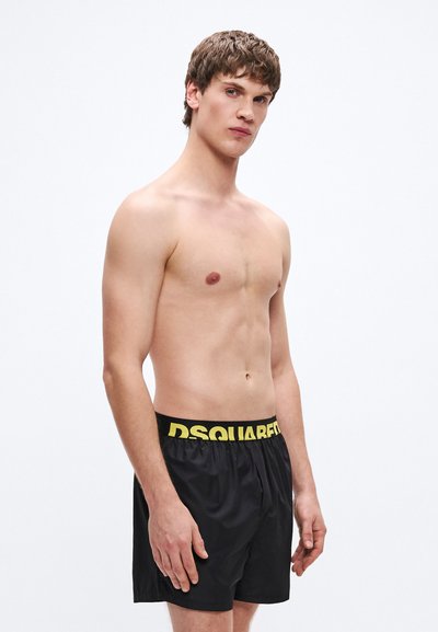 Fekete fürdőnadrág sárga "DSQUARED2" derékrésszel, könnyű anyagból, laza szabásban, semleges háttéren egy modellen bemutatva.