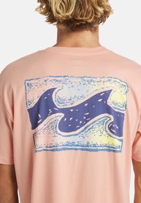 Koszulka bawełniana z krótkim rękawem w kolorze brzoskwiniowym z graficznym motywem fali w niebieskich i żółtych odcieniach na plecach. Luźny krój z gładką fakturą.