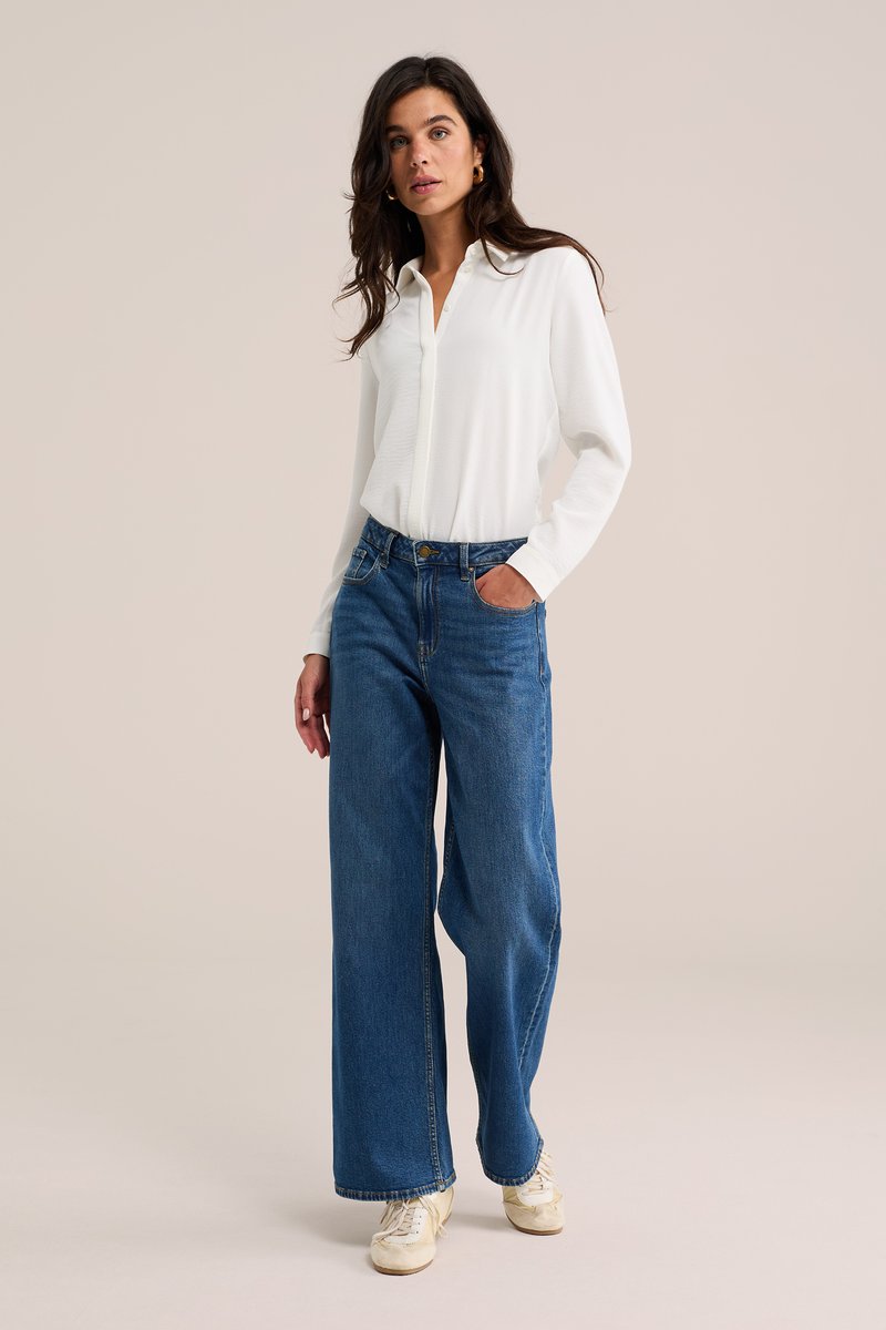 Kvinde iført en hvid skjorte med lange ærmer, blå vide jeans og hvide sneakers, der står med den ene hånd i lommen mod en ensfarvet baggrund.