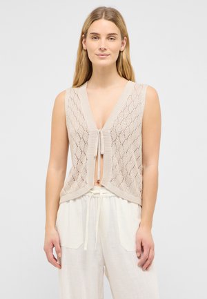 Jonge vrouw draagt een beige gebreid mouwloos vest dat vooraan is gestrikt en losse witte broek met trekkoord, staand tegen een effen achtergrond.