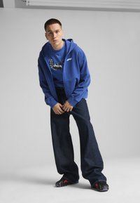 Plava dukserica na zip s plavom majicom s grafikom, u kombinaciji s širokim tamnim trapericama i crnim cipelama s crvenim akcentima.