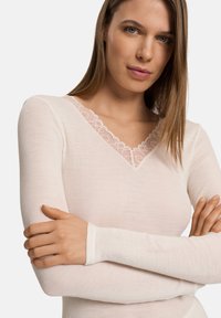 Jasnoróżowa bluzka z długim rękawem o prążkowanej teksturze i dekolcie w kształcie litery V, zdobiona kwiatową koronką. Materiał wydaje się miękki i elastyczny.