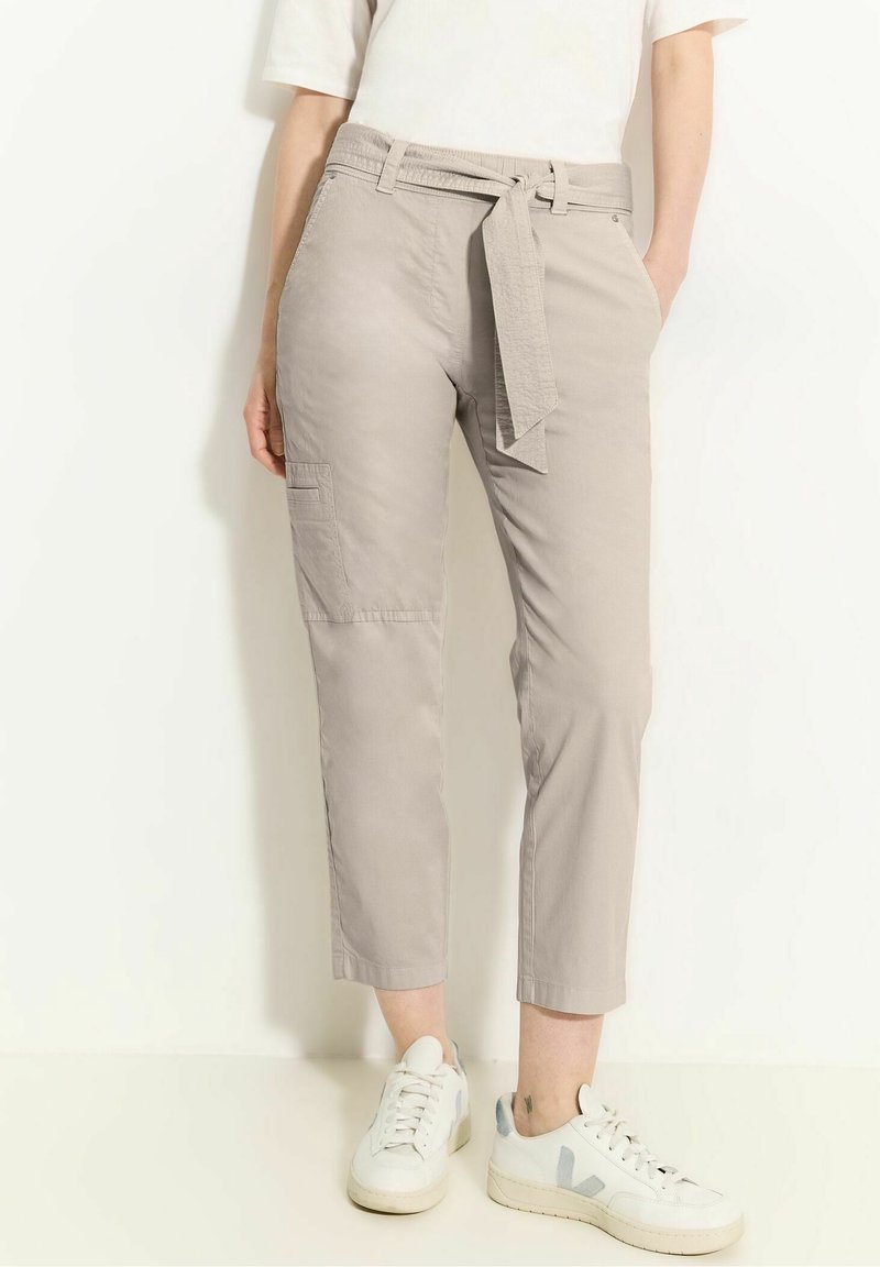 Femme portant un pantalon court beige clair avec ceinture à nouer, des baskets blanches et un haut blanc à manches courtes, debout devant un fond blanc.