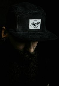 Gorra negra de pana, que cuenta con un parche rectangular blanco con el texto "Blackskies" en una fuente cursiva; la iluminación tenue resalta la textura.