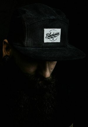 Fekete kordbársony sapka, fehér téglalap alakú folttal, rajta "Blackskies" felirattal, amely írásos betűtípussal készült; a gyenge világítás fokozza a textúrát.
