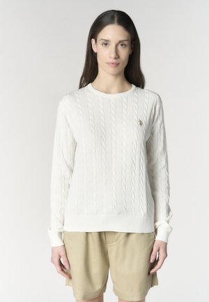Donna con capelli castani lunghi indossa un maglione bianco a trecce e pantaloncini beige, in piedi contro uno sfondo chiaro uniforme.