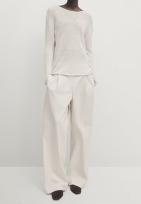 Pull en maille beige clair avec un large décolleté, associé à un pantalon ample plissé de couleur crème ; tous deux présentent des textures lisses et peu de détails.