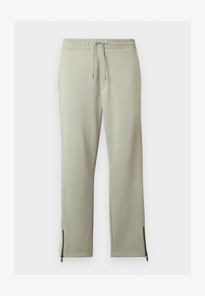 Pantalon de survêtement gris clair avec taille élastique, cordon de serrage et détails de fermeture éclair noirs aux ouvertures des chevilles, posé à plat sur fond blanc.