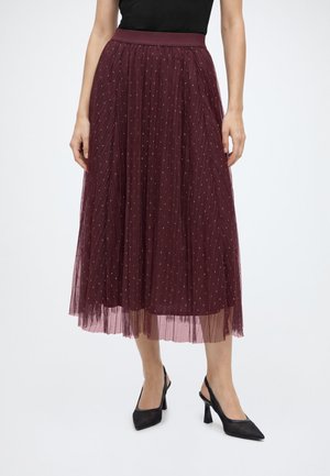 Femme portant une jupe midi plissée bordeaux avec de petits détails à pois et des escarpins slingback noirs à bout pointu.