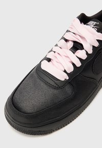 Baskets Nike Air noires avec des lacets en satin rose pâle épais et une semelle noire texturée sur fond blanc.