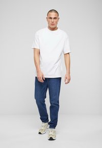 Camiseta blanca de manga corta combinada con jeans de mezclilla azules y zapatillas de color claro. El modelo está de pie frente a un fondo neutro.
