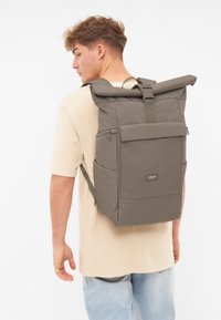 Zaino grigio a roll-top con spallacci regolabili, tasca frontale, chiusure a zip e patch con logo; realizzato in materiale resistente con una texture liscia.
