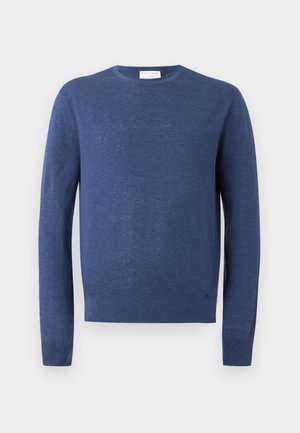 Homme portant un pull bleu à manches longues, un pantalon beige à carreaux et des chaussures noires, debout les mains dans les poches, regardant sur le côté.