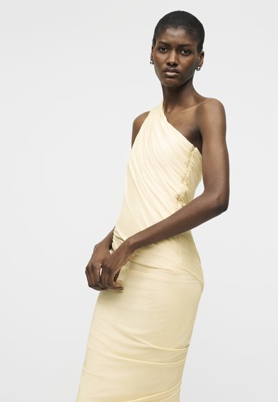 Tumšādaina sieviete, tērpusies gaiši bēšā vienas pleca rušētā kleitā, stāv ar viegli savilktām rokām vienkāršas pelēkas fona priekšā.