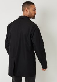 Černý kabát s límcem, vyrobený z hladkého materiálu, s rozparkem na zadní straně a dlouhými rukávy. Design je minimalistický a přiléhavý.