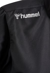 Svart sportjacka med en slät textur, med en silverfärgad "hummel"-logotyp placerad på den övre vänstra sidan nära kragen.