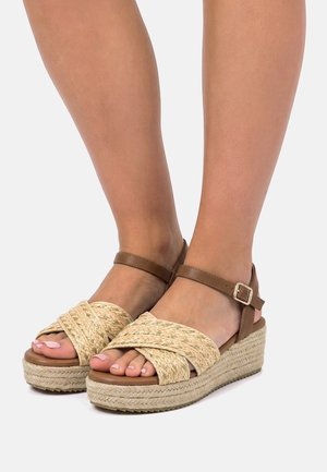 Füße, die beige gewebte Plateausandalen mit braunen Knöchelriemen und Schnallen auf einem schlichten hellen Hintergrund tragen.