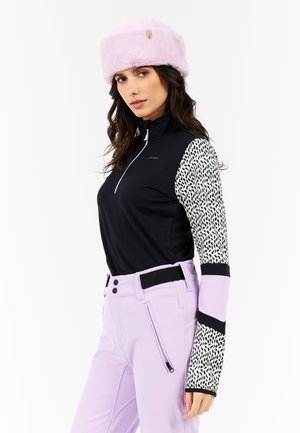Zwarte zip-up top met een patroon op de mouwen, gecombineerd met lichtpaarse broeken met riemlussen en een zijrits. Een roze bontmuts maakt de look af.