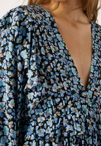 Musta kangasmekko, jossa on kukkakuosi sinisten ja keltaisten kukkien kanssa, syvä V-pääntie ja frilla-yksityiskohtia jakauksella.