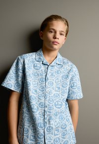 Hellblaues kurzärmeliges Hemd mit einem sich wiederholenden Muster aus Smiley-Gesichtern und Symbolen. Hergestellt aus einem leichten Stoff mit Knopfleiste vorne.