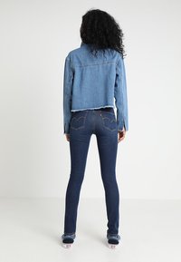 Chaqueta de mezclilla en azul claro con un diseño cropped y dobladillo deshilachado, combinada con jeans ajustados en azul oscuro que tienen bolsillos traseros y una costura visible.