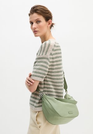 Femme aux cheveux courts portant un pull rayé vert et beige et un pantalon beige, portant un sac à bandoulière vert clair.