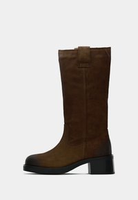 Botte haute en daim marron avec un bout rond et un talon bloc. Présente des coutures verticales et une tirette en haut pour un enfilage facile.