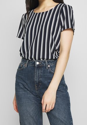 Femme portant un chemisier à manches courtes rayé verticalement noir et blanc, rentré dans un jean en denim bleu taille haute.