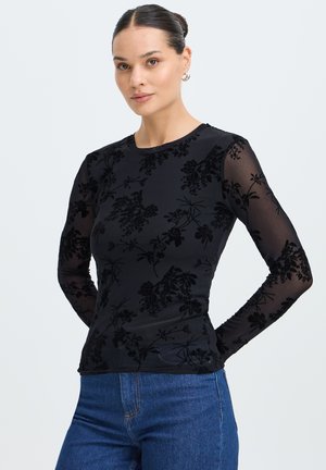 Vrouw met donker haar in een knot, die een zwarte bloemenprint doorschijnende top met lange mouwen en een blauwe spijkerbroek draagt, staand met haar handen achter haar rug.