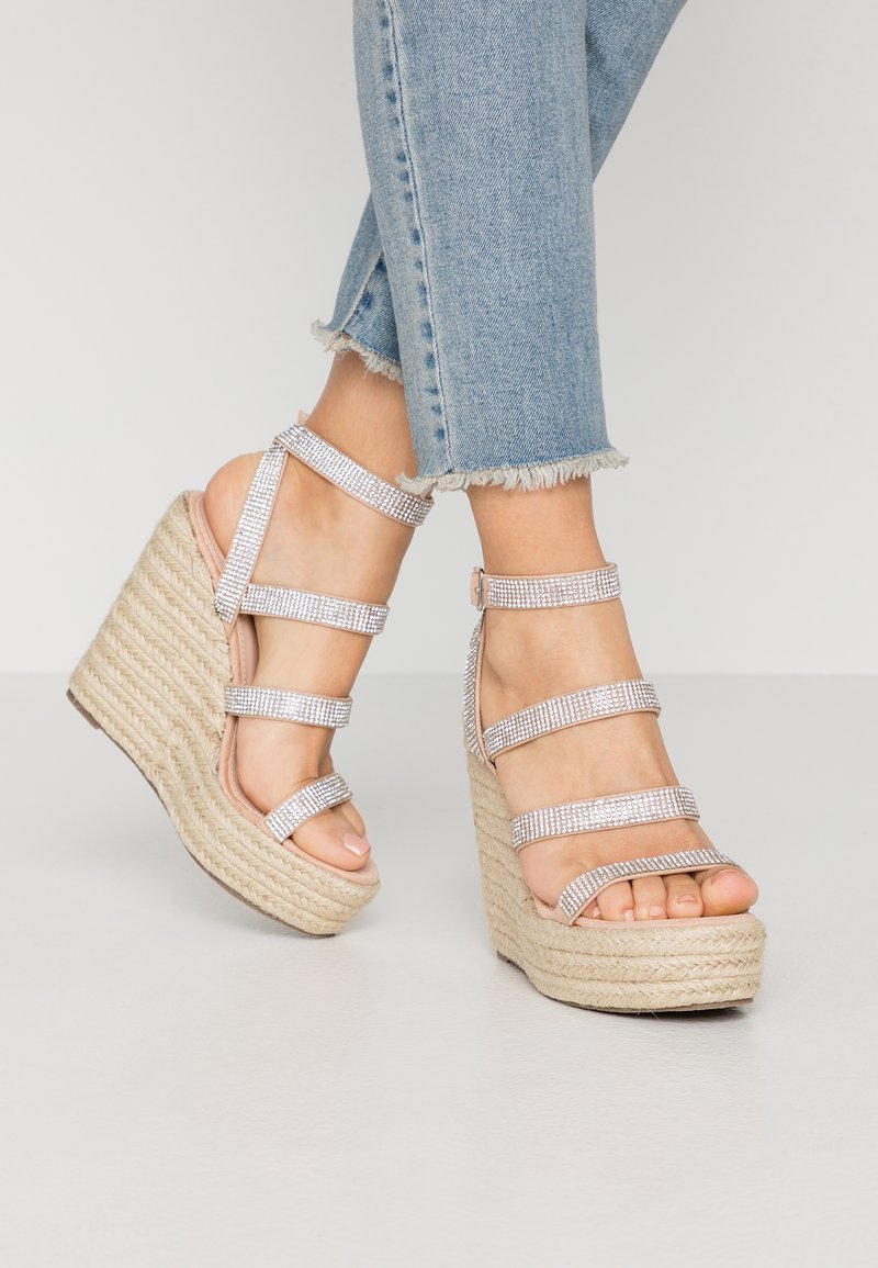 Pieds portant un jean bleu clair à ourlet effiloché et des sandales à talons compensés beiges avec des brides en strass scintillantes sur un fond clair.