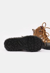 Bota de senderismo marrón con suela negra gruesa, exhibida de lado, mostrando un patrón de suela resistente y parte superior con cordones y ojales de metal, además de una cremallera lateral.