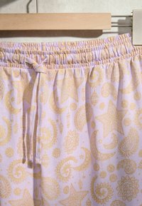 Shorts en tissu lilas avec taille élastique et lien, présentant des motifs célestes jaunes et paisley, suspendus à un cintre en bois.