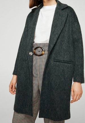 Femme portant un manteau en laine texturée vert foncé sur une chemise blanche et un pantalon taille haute à carreaux avec une boucle de ceinture ovale en écaille de tortue.