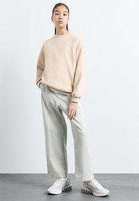 Beige oversize mikina s bílým logem, spárovaná s světle zelenými širokými kalhotami a béžovými teniskami s mřížkovaným designem a zaoblenou podrážkou.