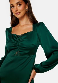 Zielona satynowa sukienka z długimi rękawami, kwadratowym dekoltem, marszczeniem na biuście oraz koronkowymi akcentami na dekolcie i u dołu. Gładka faktura.