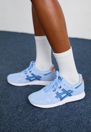 Nohy obuté do světle modrých tenisek Asics s bílými podrážkami a bílými ponožkami s vysokým střihem stojí na tmavém kobercovém podkladu.