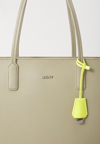 Vaaleanbeige keinokuitutote-laukku, jossa on kaksinkertaiset kahvat, kohotettu DKNY-logo ja irrotettava neonkeltaista väriä oleva etiketti. Sujuva pinta ja muotoiltu rakenne.