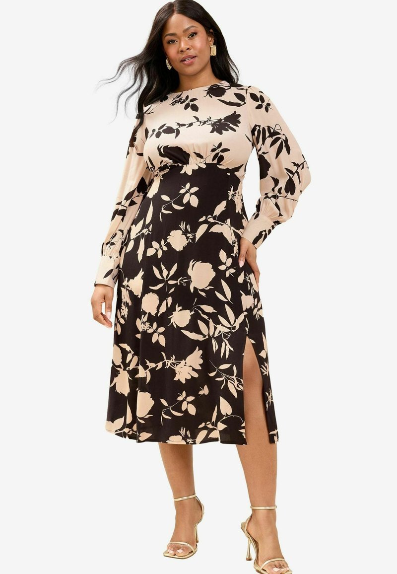 Blumengemustertes Wickelkleid aus schwarzem und beigem Stoff, mit langen Ärmeln, einem seitlichen Schlitz und einem figurbetonten Oberteil sowie einem ausgestellten Rock. Goldene Sandalen runden den Look ab.