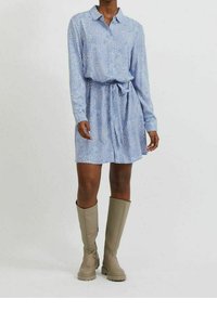 Blå, mönstrad skjortklänning med krage, långa ärmar och knytmidja. I sällskap av knähöga, beige gummistövlar.