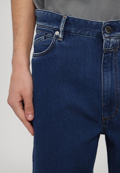 Tumed sinine teksapüksid klassikalise viie tasku disainiga, nööbi ja lukuga kinnitamine ning kontrastõmblusest detail.