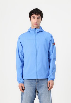 Light blue zip-up jacket with a hood, features elastic cuffs and a horizontal seam across the chest, accented with a multi-colored logo on the sleeve.
