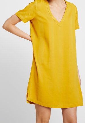 Mujer con un vestido amarillo mostaza suelto, de manga corta y escote en V, de pie con una mano en la cadera.